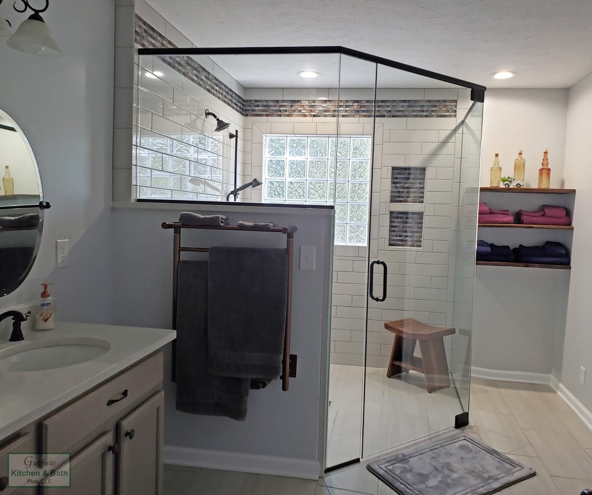 A completed Guthrie Kitchen & Bath Plus bathroom remodel in Clarksville, Tennessee: a corner frameless-glass walk-in tile shower with a bench and glass-block window, a half-wall beside a marble-topped vanity, and a tiled niche - the early layout and fixture decisions that lock a bathroom in before any finish is chosen.
