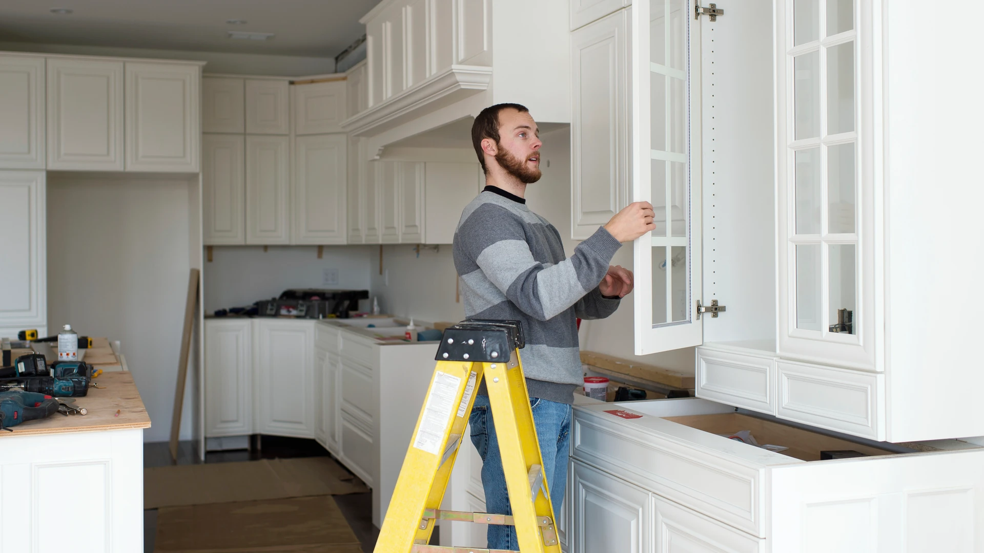 Person adjusting a glass-front upper cabinet door beside a stepladder in a white kitchen
