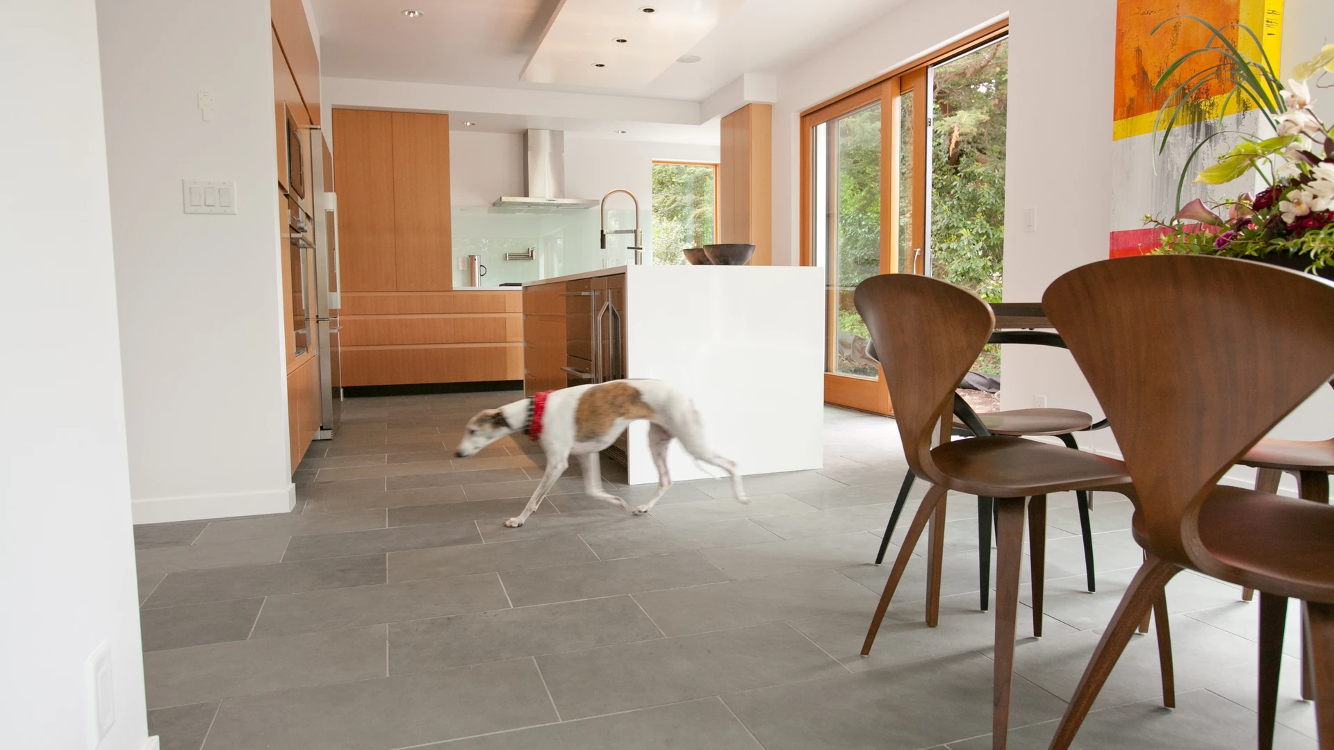 Dog trotting across a large-format gray tile floor in a modern kitchen with wood cabinets and a white island