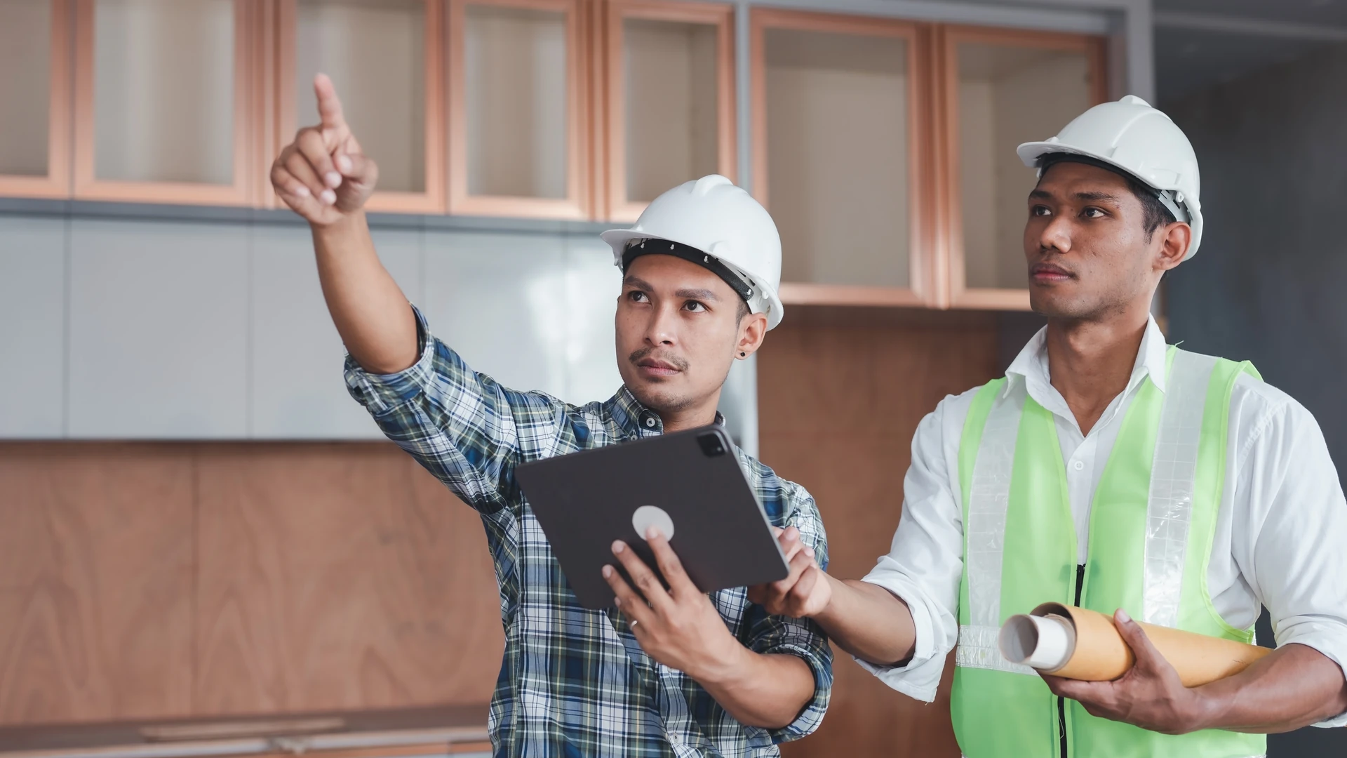 Two people in hard hats holding a tablet and rolled papers, with one pointing upward in an unfinished kitchen
