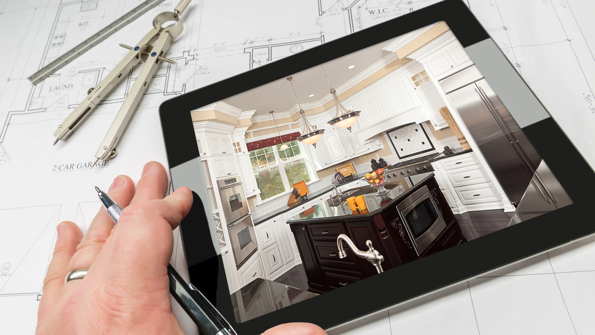 Hand with a pen beside a tablet showing a finished kitchen, resting on house floor plans with drafting tools