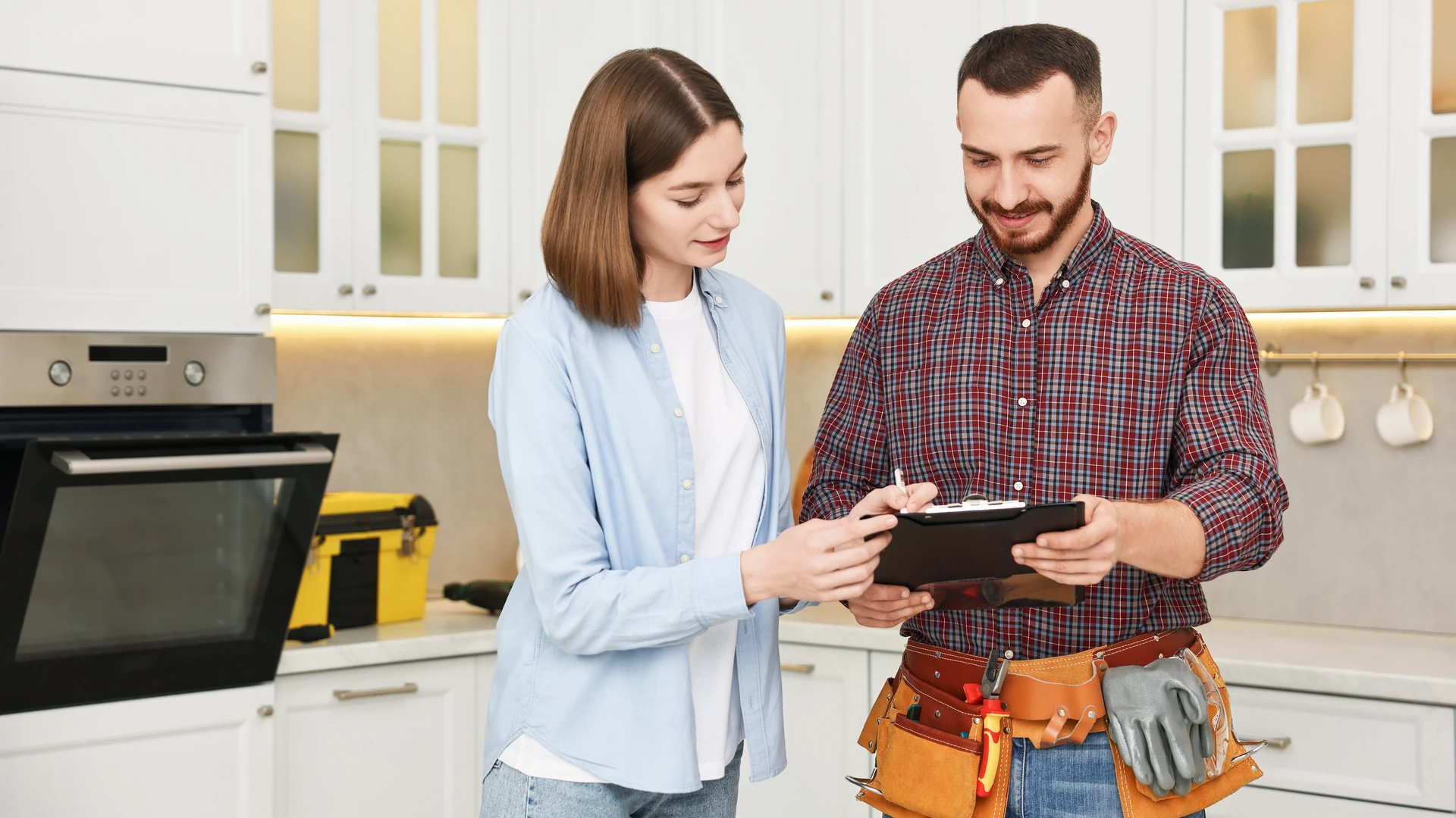 Two people reviewing a clipboard in a white kitchen, one wearing a tool belt