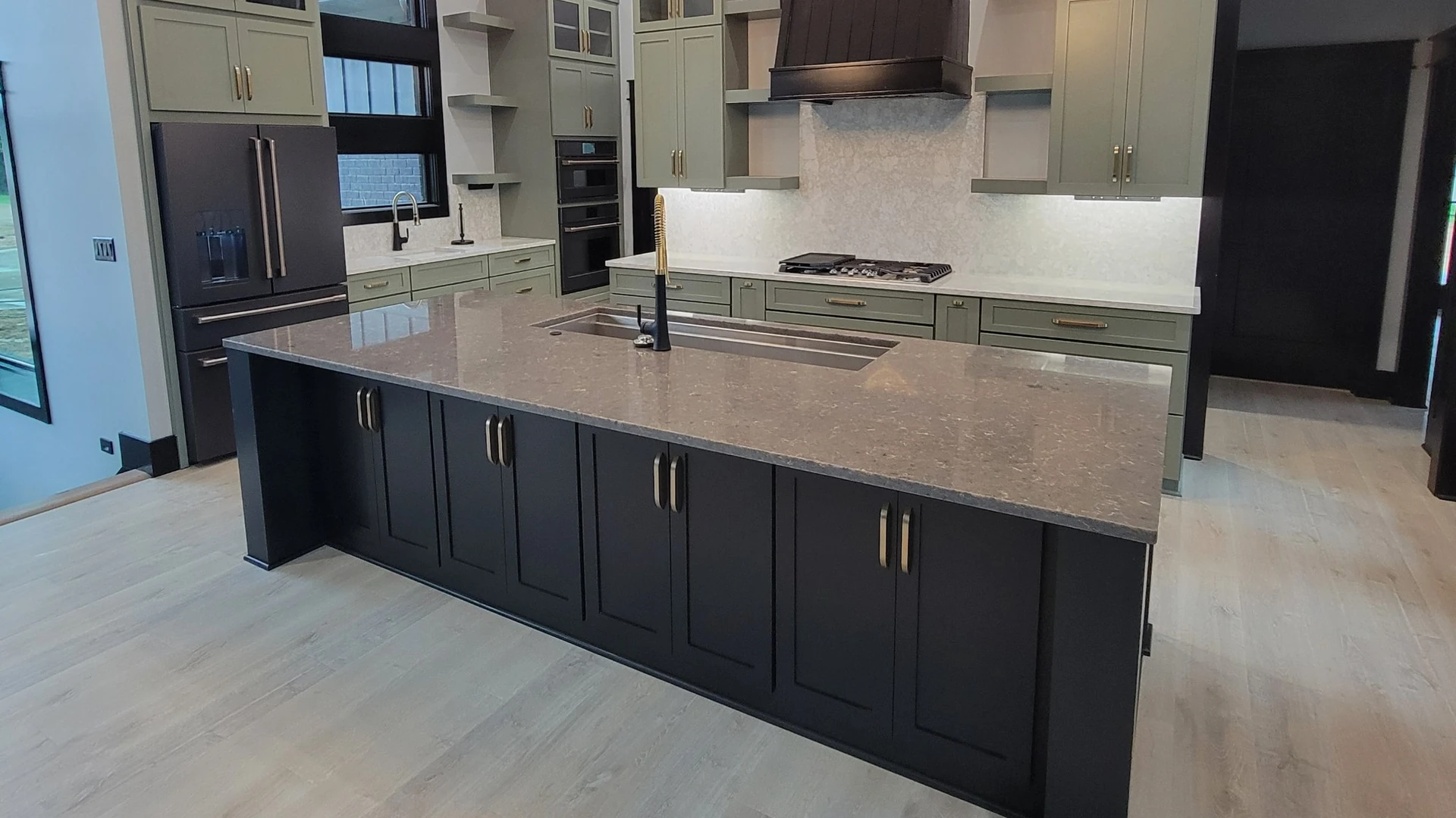 Kitchen with a dark island, sage-green cabinets, a black range hood and a light wood-look plank floor