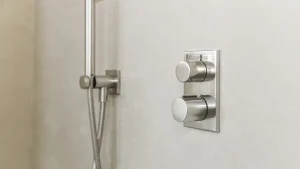 Brushed-metal wall plate with two round shower control knobs beside a vertical rail and a braided hose attached at a wall bracket