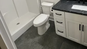 White toilet between a white shower base and a white vanity with a dark veined counter, on a dark gray tile floor