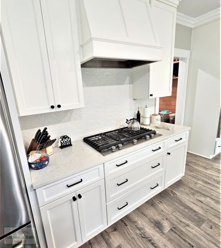 Gas cooktop beneath a white hood with a pale patterned backsplash and white cabinets